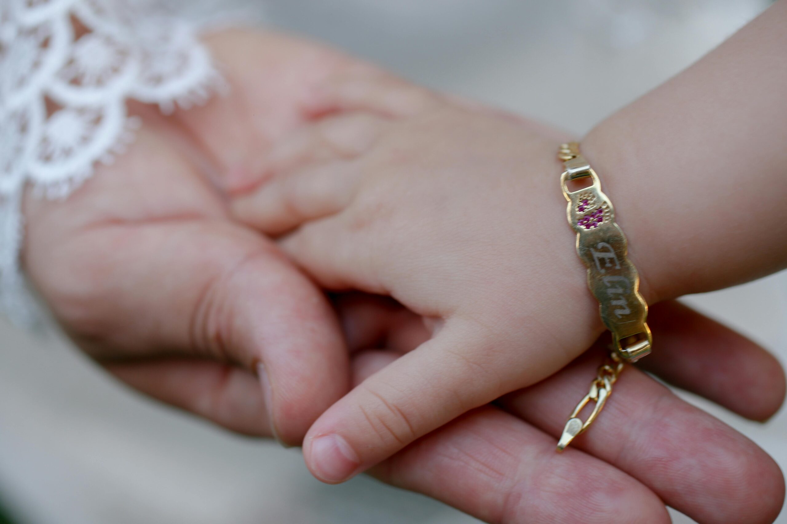 Close-up of a baby holding an adult's hand, featuring a gold bracelet with engraved name.